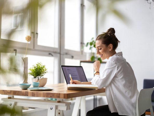 A woman sitting at a desk with a laptop. She is in front of large windows. There is a plant on the desk. She is wearing a white button up shirt and her hair is in a bun.