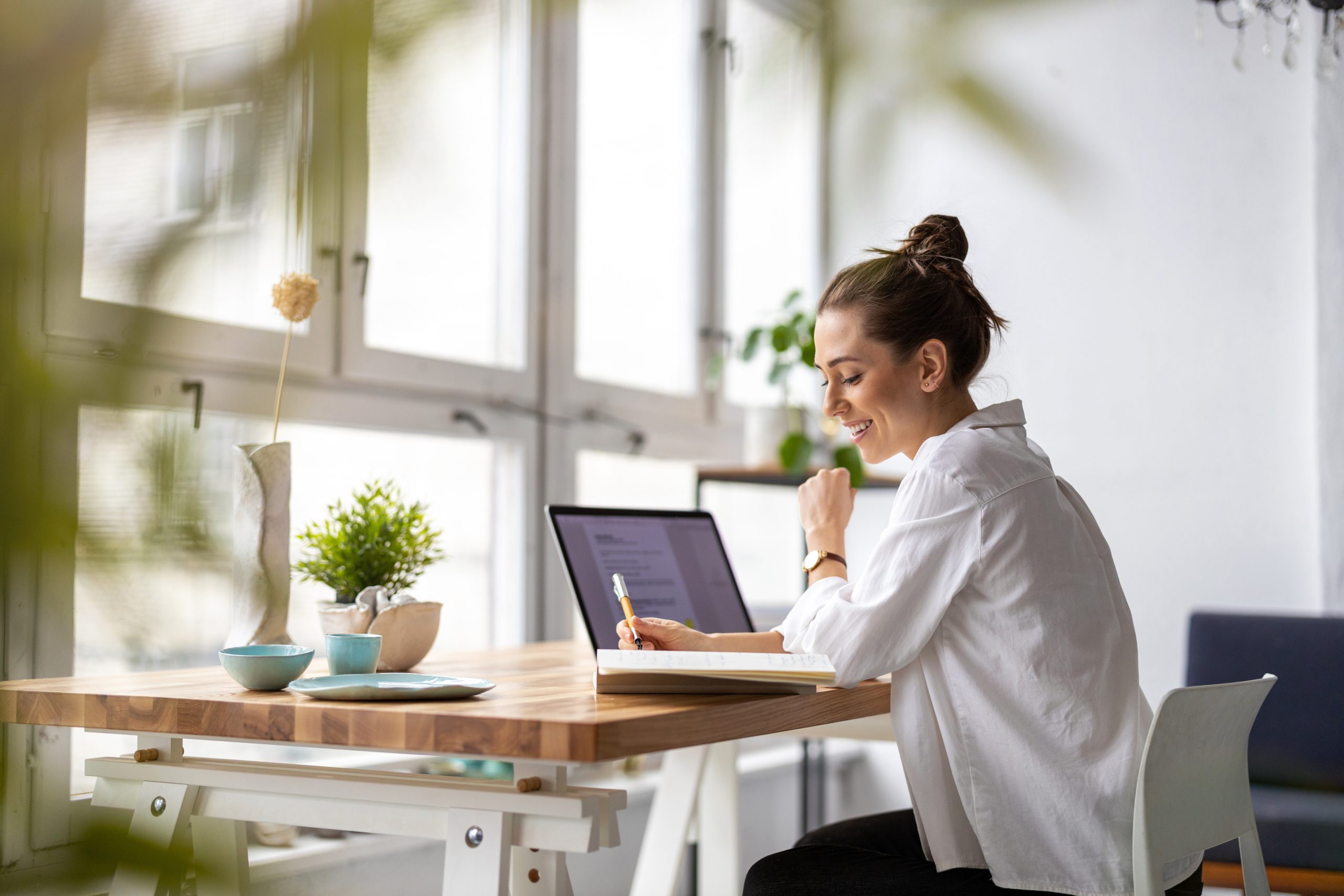 A woman sitting at a desk with a laptop. She is in front of large windows. There is a plant on the desk. She is wearing a white button up shirt and her hair is in a bun.